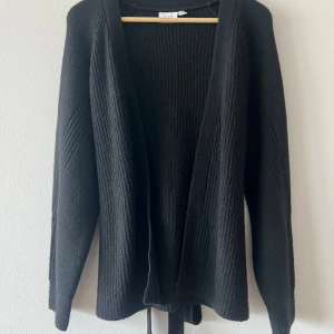 Svart stickad kofta från Vila - Svart stickad kofta från Vila med långa ärmar och snygg ribbad struktur. Koftan har öppen front och knytband i midjan för en extra cool detalj. Perfekt att slänga på sig när det är lite kyligt ute. Använd en gång