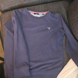 Mörkblå sweatshirt från Gant - En stilren mörkblå sweatshirt från Gant med klassisk rund hals och diskret logga på bröstet. Tröjan har långa ärmar och ribbade muddar vid ärmslut och nederkant. Perfekt för en chill och snygg look.