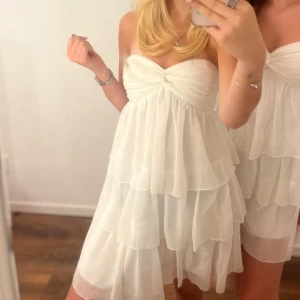 Vit axelbandslös volangklänning - Supersöt vit klänning med flera lager volanger och axelbandslös design. Klänningen har en snygg twistad detalj vid bysten och är perfekt för dig som vill ha en romantisk och luftig look. 