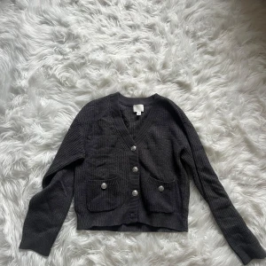 Mörkgrå stickad kofta från H&M - Säljer en mörkgrå stickad kofta från H&M i storlek XS. Koftan har v-ringning, långa ärmar och fyra silvriga knappar framtill. Två praktiska fickor med knappdetaljer ger extra stilpoäng. Perfekt att slänga på sig när det är lite kyligt ute. Knappt använd