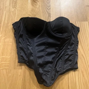 Svart satin korsett topp NAKD - Snygg svart korsett topp i satin med vadderade kupor och markerade sömmar för en riktigt cool siluett. Toppen är axelbandslös och har hysk- och hakknäppning bak. Perfekt för att lyfta vilken outfit som helst. #nakd
