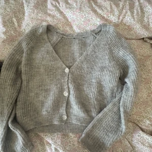 Grå ribbad kofta med v-ringning - Supermysig grå ribbad kofta med v-ringning och tre vita knappar framtill. Croppad modell med långa ärmar, perfekt att slänga över en topp eller klänning. Stickad i mjukt material som känns skönt mot huden.