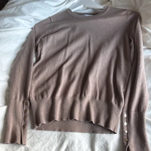Beige stickad tröja från Zara med knappar - Säljer en beige stickad tröja från Zara i storlek S. Tröjan har rund halsringning, långa ärmar och snygga metallknappar vid ärmsluten som extra detalj. Perfekt för lager-på-lager och enkel att styla till olika looks.