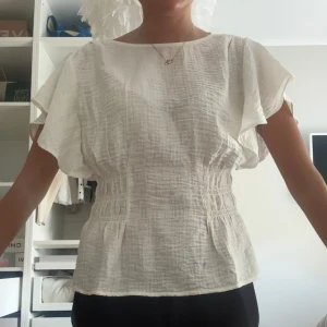 Vit blus från H&M  - Säljer en luftig vit blus från H&M i storlek XS. Blusen har korta, vida ärmar och rynkade partier i midjan som ger en snygg siluett. Tillverkad i ett lätt, strukturerat tyg som känns fräscht och somrigt. Perfekt till jeans eller kjol! Aldrig använd men hann ta bort prislappen💕