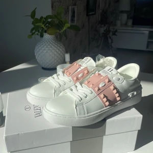 Valentino Rockstud sneakers vit/rosa - Snygga Valentino Rockstud sneakers i vitt skinn med breda rosa band och ikoniska nitar över vristen. Klassisk rund tå, platt sula och vita snören. Perfekta för dig som vill sticka ut med en lyxig och trendig look. Skorna är aldrig använda och säljs pågrund av att de var fel storlek. Pris kan diskuteras