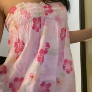 Rosa blommig axelbandslös klänning/topp - Söt axelbandslös kortklänning i ljusrosa med stora blommiga mönster i rosa och rött som kan bli vikad till en söt topp. Klänningen har smock upptill och ett luftigt, flowy fall. Perfekt för varma dagar och ger en chill vibe. Materialet känns lätt och bekvämt mot huden.