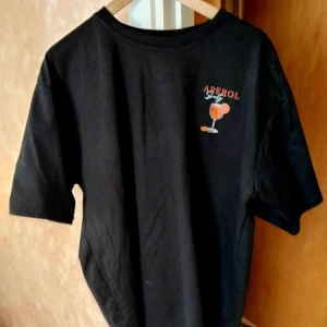 Svart Aperol Spritz t-shirt - Svart t-shirt med coolt Aperol Spritz-tryck på både bröstet och ryggen. Stor färgglad drinkillustration och text med ingredienser på baksidan. Klassisk passform och rund hals. Perfekt statement-plagg för dig som gillar snygga och roliga prints.