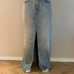 Ljusblå lång jeanskjol med slits - Säljer en lång jeanskjol i ljusblå denim med hög midja och rak passform. Kjolen har en snygg slits framtill, klassiska jeansfickor bak och en cool tvättad look. Perfekt för dig som gillar streetstyle och vill ha något trendigt i garderoben.