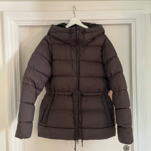 Brun pufferjacka från Everest - Säljer en brun pufferjacka från Everest med huva och dragsko i midjan. Jackan har långa ärmar, dragkedja framtill och är perfekt för kalla dagar. Fodrad och riktigt skön att bära när det är kyligt ute.