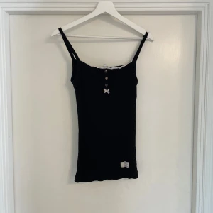 Svart ribbad linnetopp från odd molly med rosett - Svart ribbad linnetopp med smala axelband, dekorativa knappar framtill och en liten söt rosett under knapparna. Toppen har spetskant vid halsringningen och en liten etikett nertill. På baksidan finns ett broderat motiv med text och rosor.