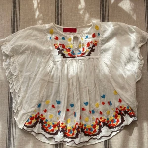 Vit blus med broderi och volangärm - Superfin vit blus i tunn bomull med färgglatt broderi i rött, blått, gult och brunt. Blusen har volangärmar, knytning vid halsen och ett luftigt, flowy fall. Perfekt för dig som gillar boho vibes och unika detaljer.