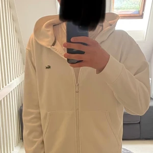 Vit hoodie från Lacoste med dragkedja - Vit hoodie från Lacoste med klassisk krokodillogga på bröstet. Tröjan har hel dragkedja, huva och två stora fickor framtill. Tillverkad i mjuk bomullsmix och har en clean, oversized look. Perfekt för en chill och stilren vibe.