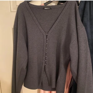 Mörkgrå kofta från Gina Tricot - Mysig mörkgrå kofta från Gina Tricot med v-ringning och dekorativa knappar framtill. Koftan har långa ärmar och ett band som kan knytas framtill. Perfekt att slänga på sig över en topp för en chill look.