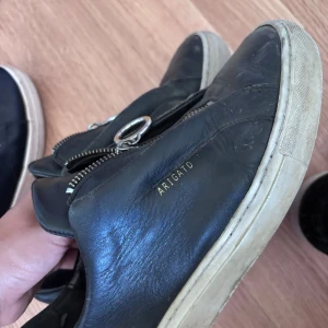 Svarta sneakers från Axel Arigato - Svarta sneakers i skinn från Axel Arigato med coola silverfärgade dragkedjor och ringdetaljer på ovansidan. Vit sula som ger en snygg kontrast. Minimalistisk design med diskret logga i guld på sidan. Perfekta för dig som gillar streetstyle med edge.