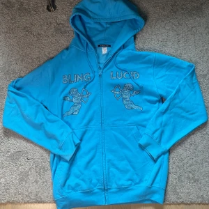 Blå hoodie från Bling Lucid - Säljer en blå hoodie från Bling Lucid med dragkedja och huva. Framsidan har coola strassdetaljer med texten 'BLING LUCID' och två änglar. Tröjan är i bomull och polyester, har känguruficka och snörning i huvan. Perfekt för dig som gillar statement-plagg.