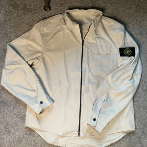 Beige overshirt från Stone Island - Snygg beige overshirt från Stone Island med klassisk patch på ärmen. Skjortan har dragkedja framtill, bröstficka och lång ärm med knapp vid manschetten. Perfekt för lager-på-lager och stilren look.