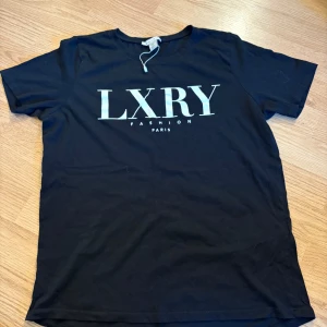 Svart LXRY t-shirt från Amisu XS - Svart t-shirt från Amisu i storlek XS med stor vit LXRY-print på bröstet och texten 'Fashion Paris' under. Klassisk rund hals och korta ärmar. Mjuk och skön bomullskvalitet, perfekt för en chill och stilren look.