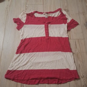 Randig topp med knappar från H&M - Säljer en kortärmad topp från H&M med breda röd-rosa och vita ränder. Toppen har rund halsringning och en knappslå framtill med fem tygklädda knappar. Tillverkad i mjuk bomull, perfekt för en avslappnad stil.