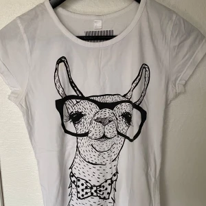 Vit t-shirt med lama från Rainbow - Cool vit t-shirt från Rainbow med ett stort print av en lama som har glasögon och fluga. T-shirten har rund hals, korta ärmar och är gjord i mjuk bomull. Perfekt för dig som gillar roliga och unika motiv.