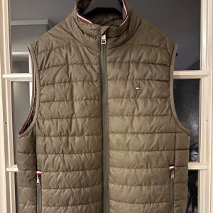 Olivgrön dunväst från Tommy Hilfiger - Snygg olivgrön dunväst från Tommy Hilfiger med quiltad design och dragkedja framtill. Västen har ståkrage och diskreta logodetaljer på bröstet och vid fickorna. Perfekt att slänga över en hoodie eller tröja för extra värme.