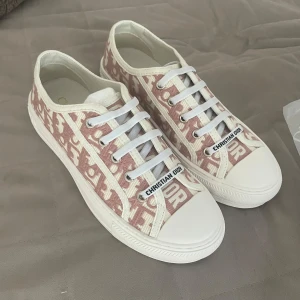 Christian Dior rosa sneakers med monogram - Snygga sneakers från Christian Dior i rosa och vitt med ikoniskt monogrammönster. Skorna har vit platt sula, rund tå och vita snören. Ovandelen är i textil med broderad logga och detaljer. Perfekta för dig som vill sticka ut med lyxig streetstyle.
