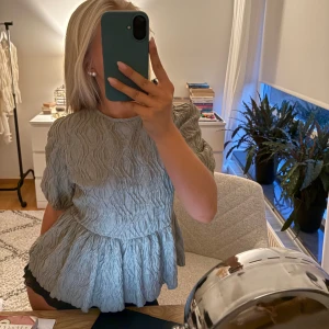 Grön volangblus från Gina Tricot - Supersöt grå blus från Gina Tricot med volang nertill och puffiga korta ärmar, har även en öppning i ryggen och knytning bak i nacken!! endast använd två gånger och i princip ny! I strl 36 🥰