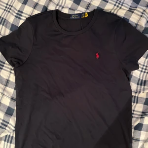 Marinblå t-shirt från Polo Ralph Lauren - Marinblå t-shirt från Polo Ralph Lauren med klassisk rund hals och korta ärmar. På bröstet sitter den ikoniska röda broderade loggan. T-shirten är gjord i mjuk bomull och har en normal passform som sitter snyggt på kroppen.