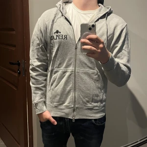 Replay hoodie - Snygg grå hoodie från Replay med svart logga på bröstet. Tröjan har huva med snörning, dragkedja framtill och två stora fickor. Tillverkad i mjuk bomull och har långa ärmar. Perfekt för en chill och avslappnad stil.