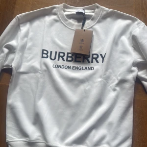 Vit sweatshirt från Burberry - Vit sweatshirt från Burberry med svart tryck 'BURBERRY LONDON ENGLAND' på bröstet. Tröjan har rund hals, ribbade muddar och är tillverkad i mjuk bomull. Perfekt för dig som gillar stilrena och klassiska designer med en lyxig touch. Helt ny men säljer för den inte passar mig. Påse ingår.