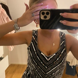 Svart paljettopp med silvriga detaljer - Säljer en svart ärmlös topp med silvriga paljetter i ett snyggt mönster som går i sicksack. Toppen  har v-ringning och är figurnära upptill. Perfekt för dig som vill sticka ut med lite extra glitter. Materialet känns festligt och skimrande.