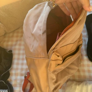 Beige ryggsäck från Giorgia & Johns - Säljer en stilren beige ryggsäck från Giorgia & Johns. Väskan har ett stort huvudfack med röd insida, en praktisk ytterficka med dragkedja och detaljer i skinnimitation. Perfekt för dig som vill ha en snygg och enkel väska till vardagen.