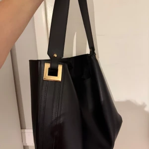 Svart axelväska med gulddetaljer - Stilren svart axelväska i slätt skinn med bred axelrem och gulddetaljer. Väskan är rymlig, har en boxig form och passar perfekt om man vill få plats med mycket. Har inte kommit till användning. 