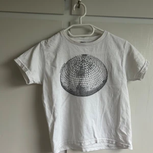 Vit t-shirt med discokula-tryck - Säljer en vit t-shirt med ett stort svart tryck av en discokula på bröstet. Klassisk passform med korta ärmar och rund hals. Perfekt för dig som gillar coola och unika prints. Materialet känns som bomull och är mjukt mot huden.