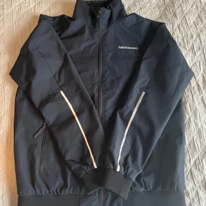 Svart Peak Performance JR coastal jacket - Säljer en svart vindjacka från Peak Performance med hel dragkedja. Jackan har ståkrage, två fickor med dragkedja och logga på bröstet. Perfekt för dig som vill ha en stilren och sportig look till hösten. 