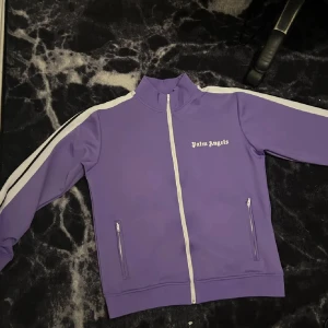 Lila Palm Angels track jacket - Säljer en lila track jacket från Palm Angels med vita ränder längs ärmarna och vit dragkedja. Jackan har ståkrage, två fickor med dragkedja och Palm Angels-logga tryckt på bröstet. Perfekt för en sportig och trendig look.