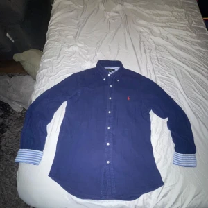 Blå skjorta Polo Ralph Lauren M - Snygg blå skjorta från Polo Ralph Lauren i slim fit med klassisk krage och knappar framtill. Skjortan har långa ärmar med blå- och vitrandiga detaljer på insidan av manschetterna samt röd broderad logga på bröstet. Perfekt för dig som gillar stilrena plagg med en twist.