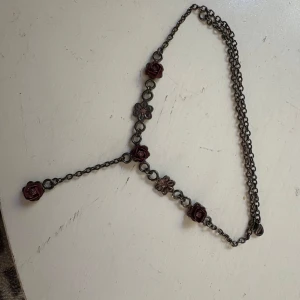 Blommigt halsband med rosor - Unikt halsband med mörk kedja och detaljerade röda rosor samt små blommor i metall. Kedjan har en Y-formad design med en ros som hänger ned. Perfekt för dig som gillar romantiska och bohemiska smycken.