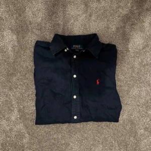 Mörkblå skjorta från Polo Ralph Lauren - Klassisk mörkblå skjorta från Polo Ralph Lauren med röd broderad logga på bröstet. Skjortan har knappar framtill och en stilren krage. Perfekt för dig som gillar en tidlös och clean look.