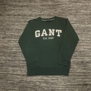 Mörkgrön sweatshirt från GANT - Snygg mörkgrön sweatshirt från GANT med vit logga och texten 'Est. 1949' på bröstet. Tröjan har rund halsringning, ribbade muddar och är långärmad. Perfekt för dig som gillar klassisk och stilren streetwear. Det står storlek 9-10y men skulle säga att den även passar upp till 11y