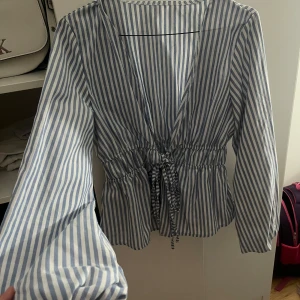 Blårandig omlottblus  - Snygg blårandig omlottblus från SHEIN i storlek S. Blusen har långa ärmar, knytning i midjan och v-ringning. Perfekt för dig som gillar en fräsch och luftig stil. Materialet är lätt och känns skönt mot huden.