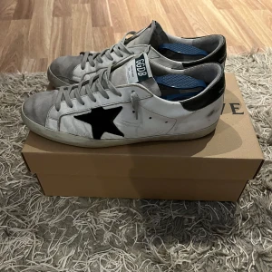 Golden Goose superstar vita sneakers med svart stjärna - Golden Goose Superstar sneakers i vitt skinn med grå mockadetaljer och svart stjärna på sidan. Skorna har svarta hälpartier, snörning och platt sula. Klassisk streetstil med GGDB-logga på plösen och hälen. Perfekta för dig som gillar en avslappnad och trendig look. OBS originalboxen tillkommer ej! Bara att skriva vid funderingar!