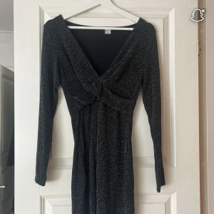Svart glittrig kortklänning med v-ringning - Säljer en svart glittrig kortklänning med lång ärm och djup v-ringning framtill. Klänningen har en twistad detalj vid bysten och är figurnära i modellen. Perfekt för dig som vill sticka ut med lite extra glitter.