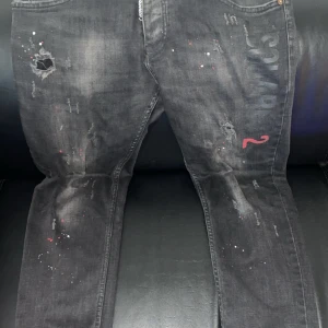 Svarta Dsquared2 jeans med tryck - Säljer ett par svarta jeans från Dsquared2 med slitningar, färgstänk och tryckt logga på ena benet. Jeansen har en röd detalj och patch bak i midjan. Snyggt street-inspirerat stuk och smal passform. Perfekta för dig som gillar en edgy look.