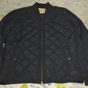 Mörkblå quiltad bomberjacka Holly & Whyte - Snygg mörkblå quiltad bomberjacka från Holly & Whyte by Lindex. Jackan har dragkedja framtill, ribbade muddar och två fickor med tryckknappar. Perfekt för dig som gillar stilren och sportig look.