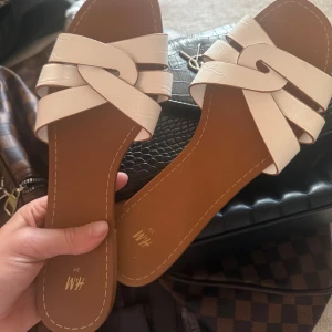 Vita sandaler med flätad rem från H&M - Snygga vita sandaler från H&M med flätad remdesign och platt sula. De har en stilren look med breda remmar i syntetmaterial och en brun innersula. Perfekta för varma dagar och enkel att matcha med sommarens outfits. Oanvända