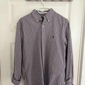 Rutig skjorta från Lauren, lila/grön - Snygg rutig skjorta från Lauren i lila, grön och vit. Skjortan har klassisk krage med knappar, lång ärm och normal passform. Perfekt för dig som gillar stilrena och färgglada plagg. Bomullsmaterial som känns skönt mot huden. Custom fit, passar M. Endast använd några få gånger