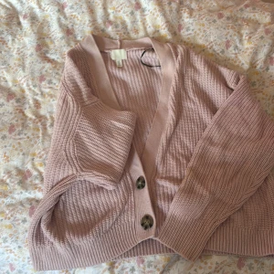 Rosa stickad kofta från H&M - Mysig ljusrosa stickad kofta från H&M med stora knappar framtill. Koftan har ribbade muddar och en relaxed passform, perfekt att slänga på sig över en t-shirt. Materialet är mjukt och känns skönt mot huden.