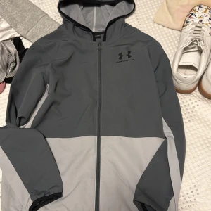 Under Armour vindjacka grå/svart - Snygg tvåfärgad vindjacka från Under Armour i mörkgrått och ljusgrått. Jackan har huva, meshfoder och dragkedja framtill. Perfekt för blåsiga dagar och sportiga outfits. Logga på bröstet och elastiska muddar vid ärmslut. Lite för liten för mig som är 170