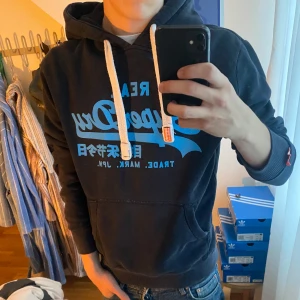 Mörkblå Superdry hoodie med tryck - Säljer en mörkblå hoodie från Superdry med stort blått tryck på bröstet och japanska tecken. Tröjan har vit snörning i huvan, känguruficka och ribbade muddar. Perfekt för dig som gillar streetwear och vill ha en snygg och bekväm look.