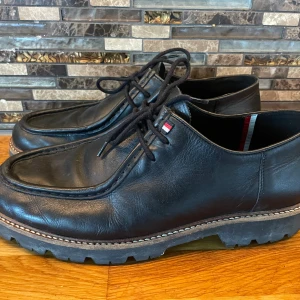 Svarta skinnskor Tommy Hilfiger - Klassiska svarta skor från Tommy Hilfiger i skinn med grov sula och snörning. Skorna har rund tå och diskret logodetalj i rött, vitt och blått vid plösen. Perfekta för en stilren och avslappnad look.